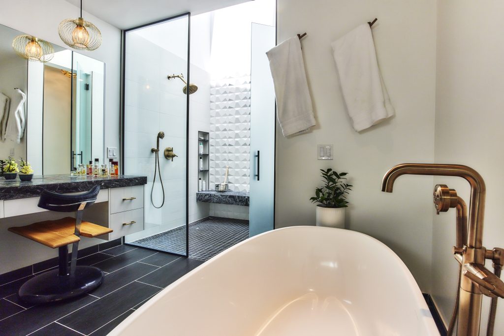 A modern bathroom featuring a large bathtub with a bronze faucet in the foreground, a shower area with glass doors and white tile walls, a vanity with a wooden chair, and hanging pendant lights. Two white towels hang on a hook beside a green plant.