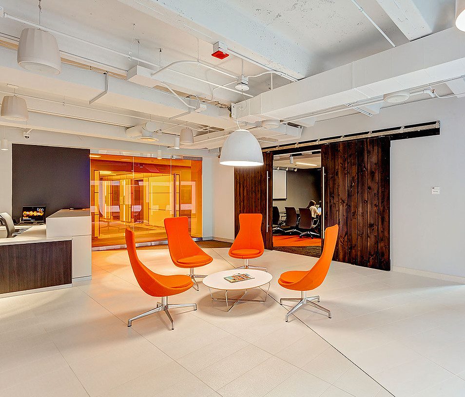 A modern office space featuring a circular arrangement of four orange chairs around a white round coffee table. Behind them is a glass-walled conference room with an orange interior. The room has white walls, a white ceiling with exposed ducts, and sleek, minimalist decor.