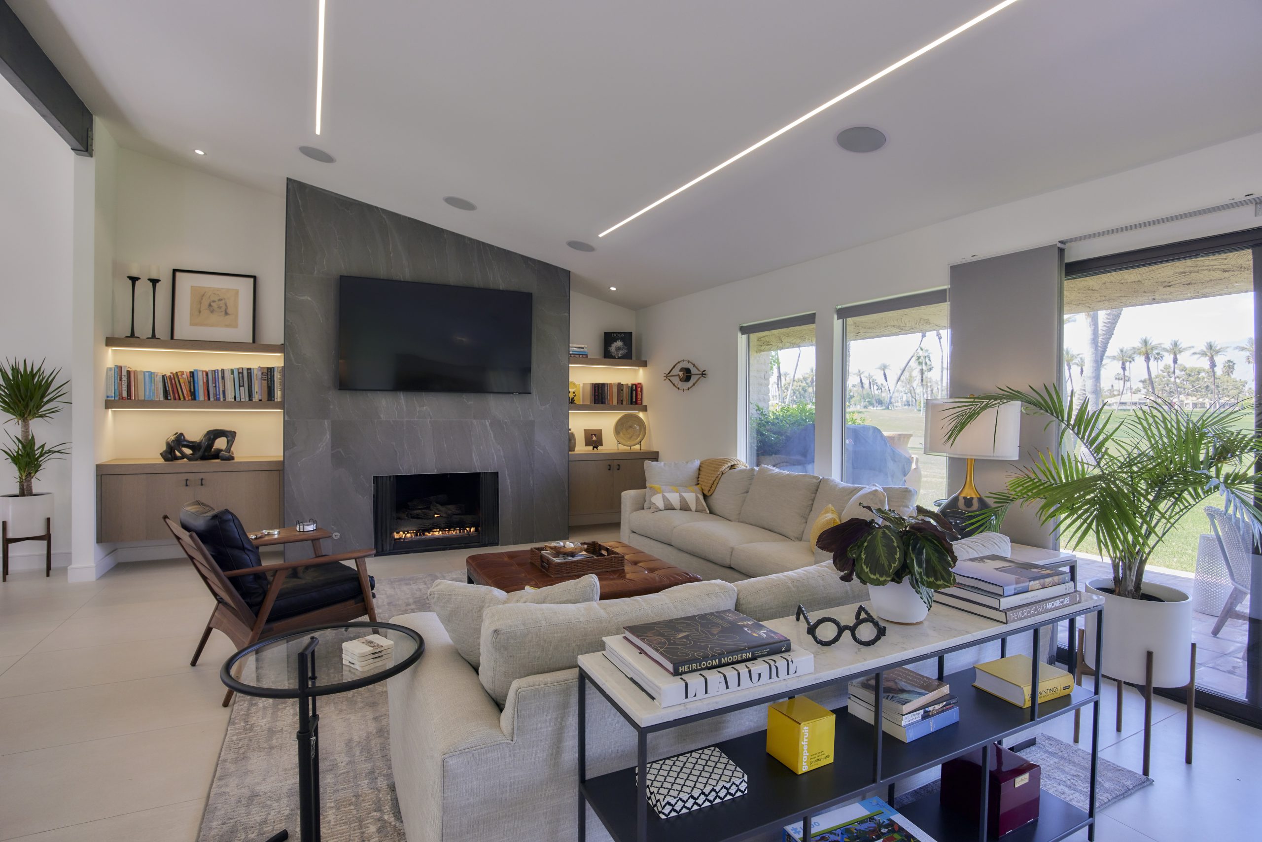 A modern living room featuring a large sectional sofa, armchairs, and a coffee table. There's a fireplace beneath a mounted TV, surrounded by built-in bookshelves. Large windows with views of greenery offer plenty of natural light. Indoor plants add a fresh touch.