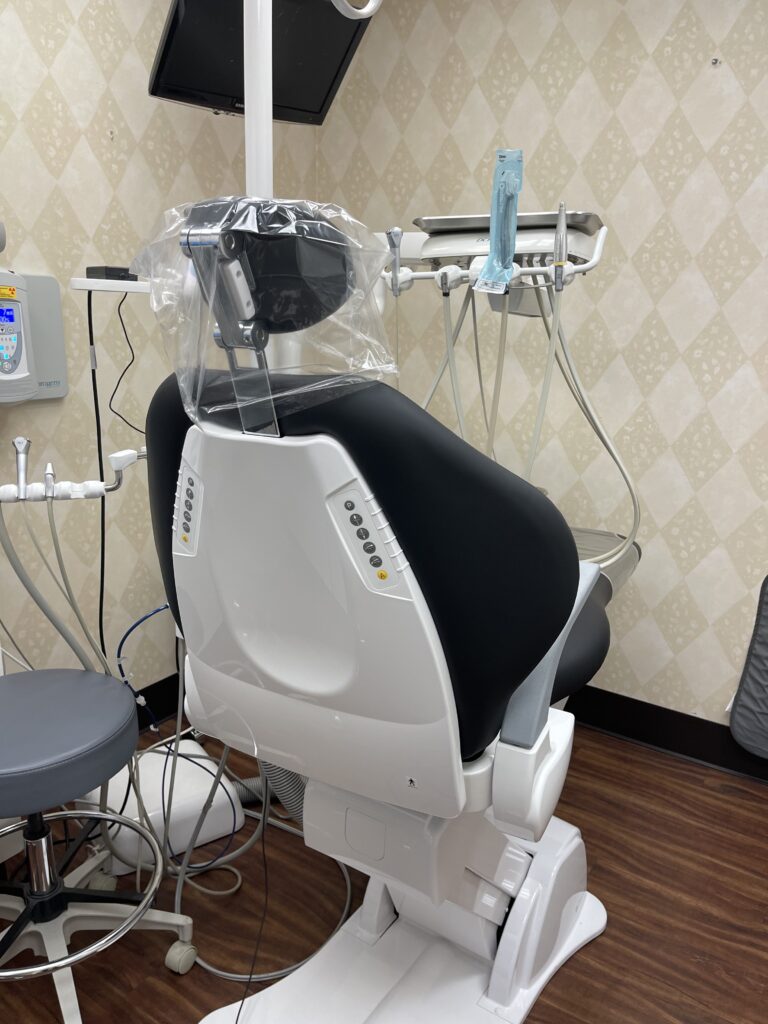 A dental exam chair with a plastic cover over the headrest, surrounded by dental equipment and a stool.