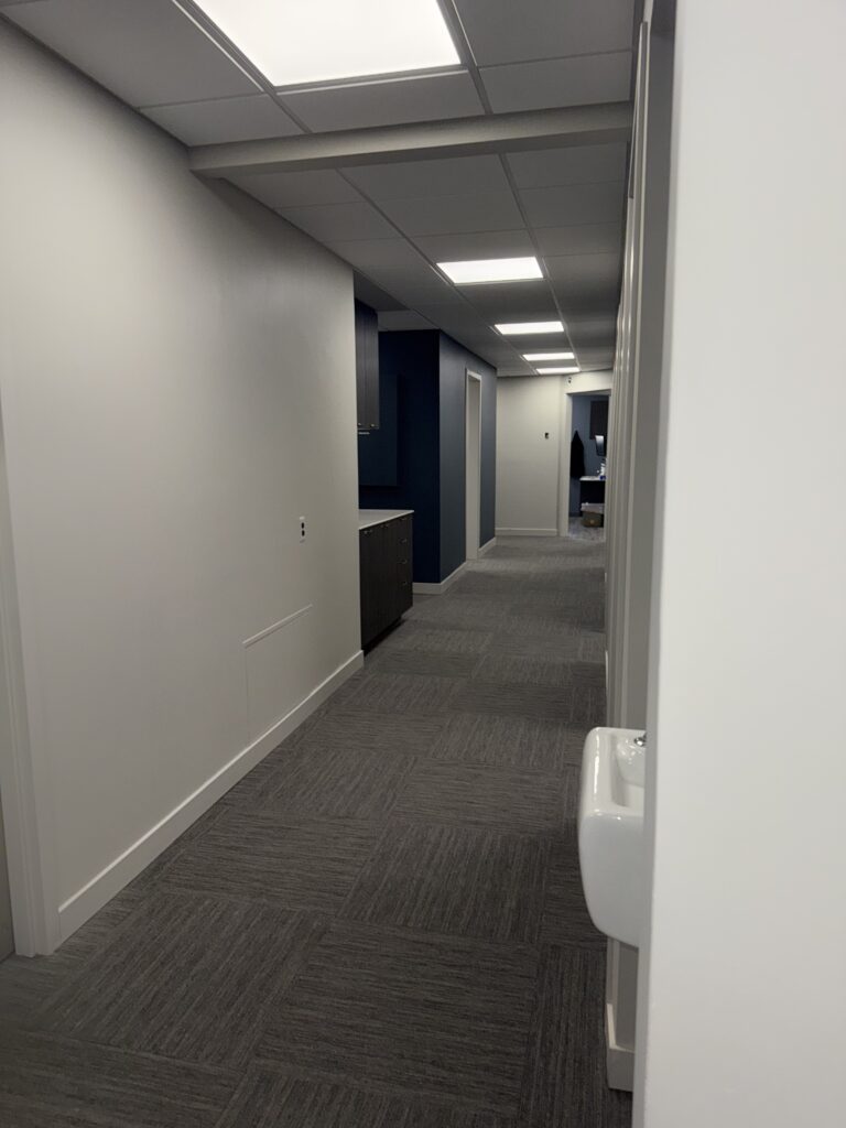 A long office hallway with gray carpet, white walls, and a small dark blue alcove with cabinetry.