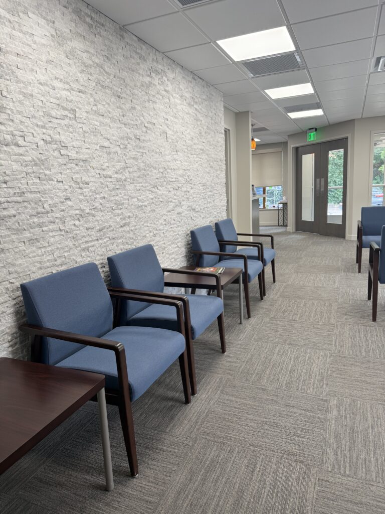 Waiting area with a row of blue chairs along a textured stone wall, with a hallway leading to an exit door in the background.