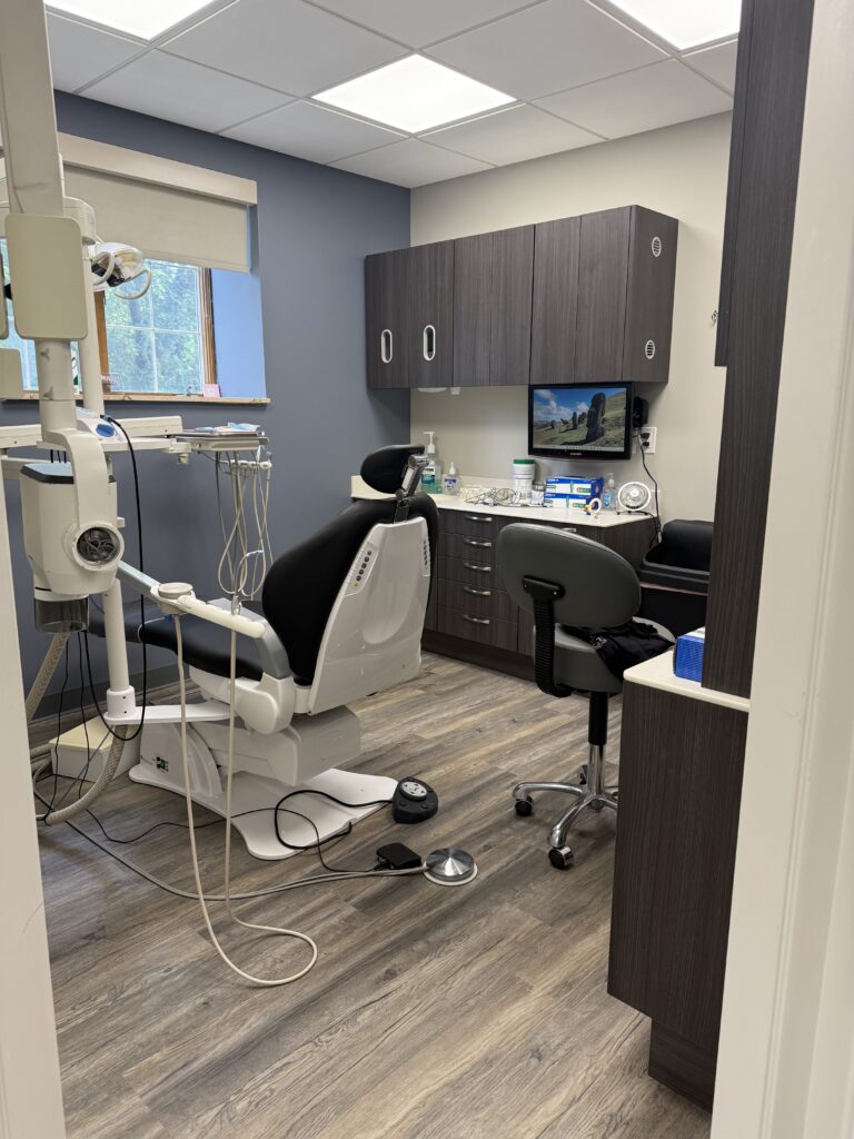 A dental examination room featuring a patient chair, a stool, dental equipment, cabinetry, and a wall-mounted monitor displaying a scenic landscape.