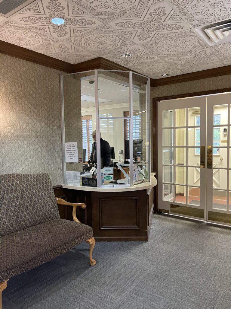 A waiting area with a patterned sofa facing a glass-enclosed reception desk, where a staff member sits behind a sign reading “We make every effort to see you on time.”